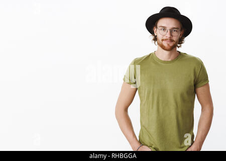 Taille plan sur beau mec optimiste avec chapeau, lunettes avec tatouages, piercing et barbe tenant les mains dans les poches en souriant joyeusement, dégustez une cuisine Banque D'Images