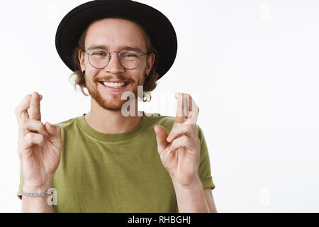 Jusqu'à la taille de balle heureux et sincère genre jeune homme barbu traveler décisions espère ou avec les mains et les doigts croisés, souriant à optimiste Banque D'Images