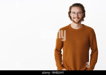 Taille plan sur beau charmant barbu à lunettes avec la longue chevelure ondulée en sweater smiling at camera joyeusement comme détenant les mains dans les poches et Banque D'Images