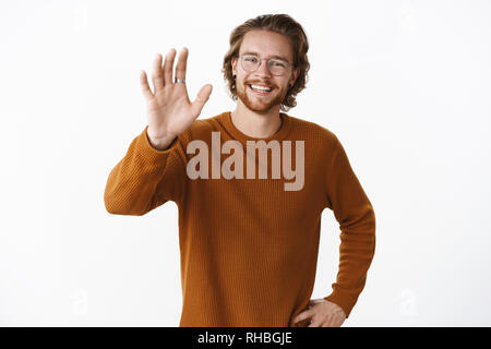 Studio shot of ordinary friendly et heureux jeune homme barbu à lunettes et pull forme main levée en geste bonjour smiling at camera comme message d'accueil Banque D'Images