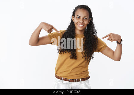 Charmant friendly-à genre et timide africains-américains femme féminine avec de longs cheveux bouclés en flexion pointant sur elle-même comme le bénévolat, smiling Banque D'Images