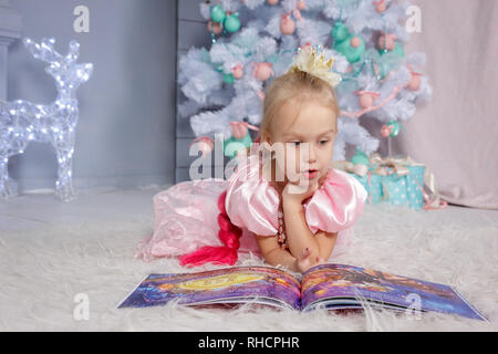 Portrait of cute little girl européenne blonde princesse avec couronne en belle robe gisant sur le sol et la lecture réserve en studio décoré dans christm Banque D'Images