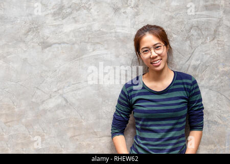 Jolie jeune femme asiatique avec des lunettes en souriant mur de béton gris Banque D'Images