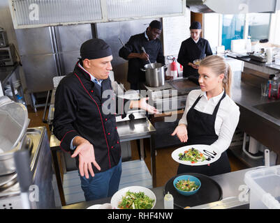 Jeune serveuse debout avec plat cuisiné dans une cuisine de restaurant à parler avec perplexe male chef Banque D'Images