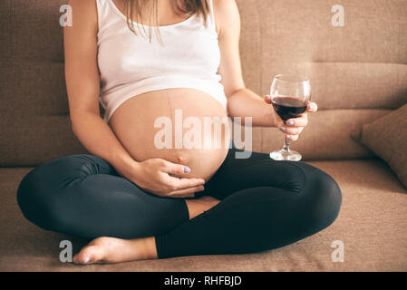 Portrait de la future mère en chemise blanche et pantalon noir assis sur le canapé à la maison et de boire de l'alcool. Femme enceinte de boire du vin pendant la grossesse période. Concept de menace, de vie malsain. Banque D'Images