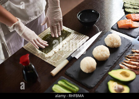 Close-up view of chef's hands préparer sushi roulant avec des ingrédients : riz, nori, avocat, saumon, sauce soja Banque D'Images