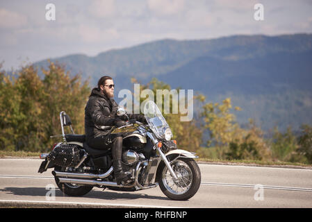 Vue latérale d'un motocycliste à poil long barbu à lunettes de soleil et des vêtements de cuir noir moto cruiser équitation le long chemin d'asphalte étroit aux beaux jours Banque D'Images