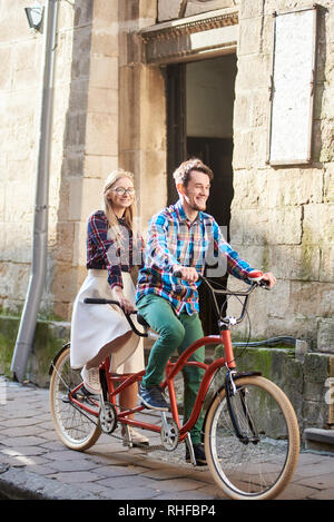 Happy tourist couple actif, barbu et souriant femme blonde aux cheveux longs en jupe blanche et verres vélo tandem le long des trottoirs de la ville pavée sur journée ensoleillée par de vieux bâtiment en pierre ancienne. Banque D'Images