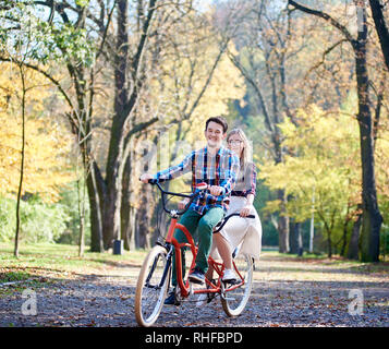 Jeune couple actif, beau barbu et attractive blonde long-haired woman vélo tandem vélo double ensemble par l'allée ensoleillée avec des feuilles d'or sur fond de grands arbres Banque D'Images