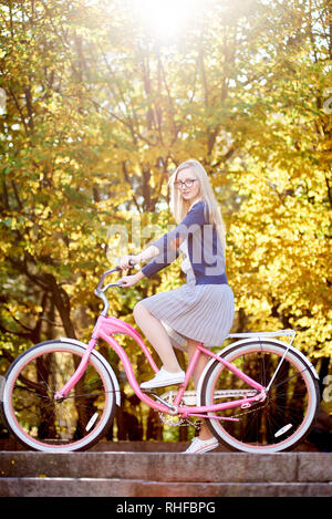 Belle blonde aux cheveux longs fille touristiques dans les verres, jupe et un chemisier qui posent sur la modernisation de la dame rose location sur allumé par autumn sun park escaliers sur bright colorful bokeh background arbres d'or. Banque D'Images