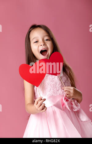 Cute little girl rire dans une robe rose tenant deux papier sur fond rose. La Saint-Valentin Banque D'Images