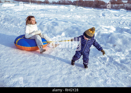 Petit garçon enfant tire sur la corde. En salopette bleue et un chapeau. Femme maman est assise sur un jouet. Heureux de jouer en hiver à l'extérieur souriant. Banque D'Images