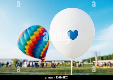 Ballon hélium blanc avec un coeur plus grande foule floue autour de montgolfières multicolores prêts à s'envoler dans un ciel bleu clair Banque D'Images