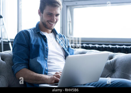 Young attractive smiling guy consulte à son ordinateur portable, assis à la maison sur le canapé gris à la maison, le port de tenue décontractée. Banque D'Images