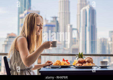 Une jeune femme est de prendre le petit déjeuner sur le balcon. Table de petit-déjeuner avec café et fruits pain croisant sur un balcon sur la toile de la grande Banque D'Images