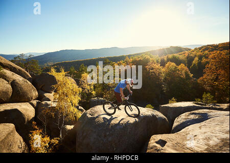 L'équilibrage de l'athlète cycliste masculin en procès location, ce truc acrobatique au-dessus de big rock le soir d'été, ciel bleu, forêt et le coucher du soleil sur l'arrière-plan. Concept de sport extrême de vie actif Banque D'Images