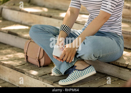 Jeune femme en bleu jeans et baskets à rayures se trouve sur d'anciennes marches de bois et est titulaire d'un téléphone dans ses mains. Focus sélectif. Banque D'Images