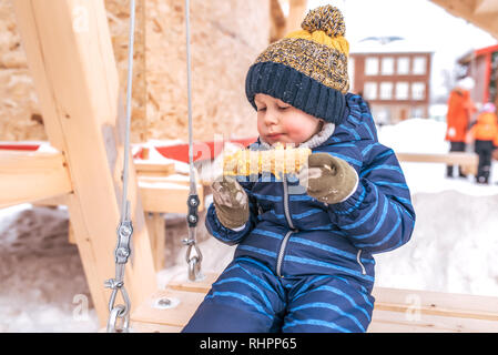 Petit garçon enfant 2-6 ans, manger du blé en ville dans l'hiver à l'air frais dans une salopette bleue chapeau et mitaines. Équitation sur une balançoire en bois. Gratuitement Banque D'Images