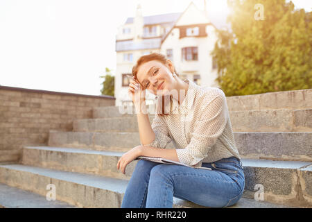 Belle jeune élève écrit un essai dans son portable assis sur les marches des escaliers piscine fille rousse avec des taches de rousseur étudier à temps libre Banque D'Images