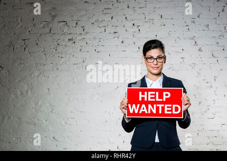 Portrait of beautiful young businesswoman holding help wanted sign against brick wall Banque D'Images