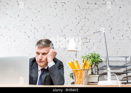 Ennuyer mature businessman looking at laptop sur 24 contre brick wall in office Banque D'Images