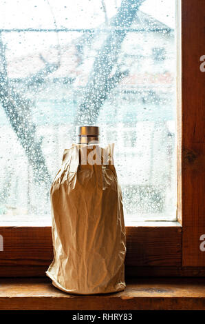 Bouteille avec de l'alcool dans un pack brown papier placé à la fenêtre au jour de pluie. L'emballage du papier froissé. Bouteille cachés de l'alcool en papier. Concept pour voler Banque D'Images