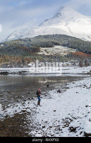 Femme sur les rives du Loch Leven lors d'une froide journée d'hiver enneigée avec pap of Glencoe, Sgorr na Ciche, village de Glencoe, Highlands, Scotland Banque D'Images