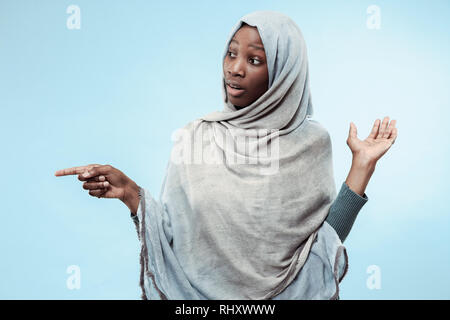 La belle jeune fille musulmane d'Afrique noire portant le hijab gris bleu au studio. Elle pointe vers la gauche et debout avec un sourire heureux sur son visage. Les émotions humaines, l'expression faciale concept. Couleurs tendance Banque D'Images