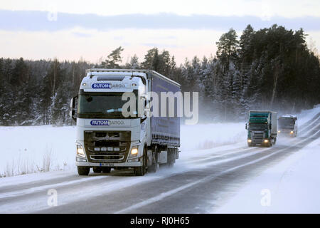 Salo, Finlande - le 18 janvier 2019 : Blanc Volvo FH d'Rutomcargo, Allemagne, vert et d'un camion Scania bus voyage à travers des paysages d'hiver finlandais au crépuscule. Banque D'Images