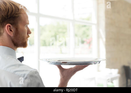 Vue arrière du confident young waiter holding tray debout au restaurant Banque D'Images