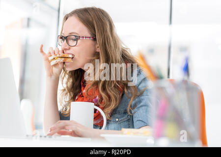 Hungry woman eating sandwich while using laptop Banque D'Images