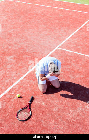 Toute la longueur de déçu man with head in hands tout en s'agenouillant par raquette de tennis sur cour rouge pendant l'été Banque D'Images