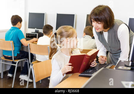 Friendly woman paying tween girl durant la leçon dans la salle informatique de la bibliothèque de l'école Banque D'Images