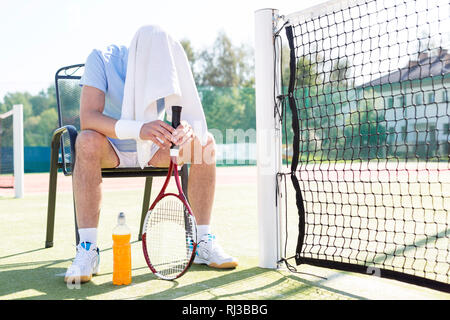 La longueur totale de l'homme mature fatigué avec la tête couverte assis sur une chaise en filet au tennis sur sunny day Banque D'Images