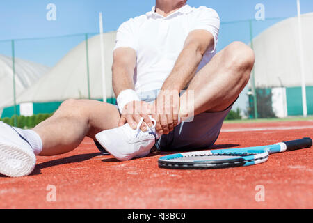 La section basse de l'homme mûr qui s'étend de la jambe en position assise sur le rouge de tennis en été Banque D'Images