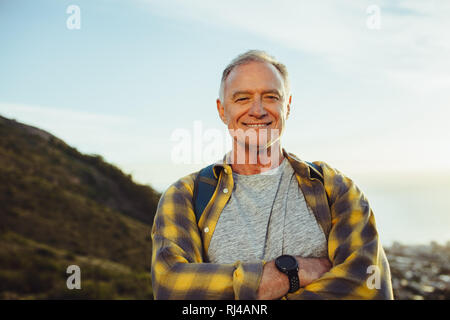 Portrait of a cheerful senior homme debout à l'extérieur avec les bras croisés. Smiling tourist homme portant un sac à dos debout sur une colline. Banque D'Images