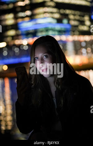 Le visage d'une adolescente est éclairé par son smartphone avec l'horizon de la ville lumières soufflées dans la distance. Banque D'Images