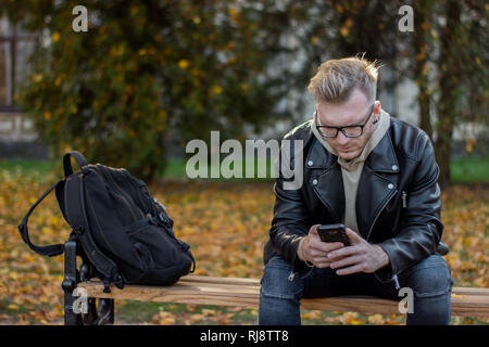 Rêver smiling guy avec un téléphone dans les tenues, assis sur le banc, la réussite d'un élève en lunettes noires et une veste à profiter de la vie Banque D'Images