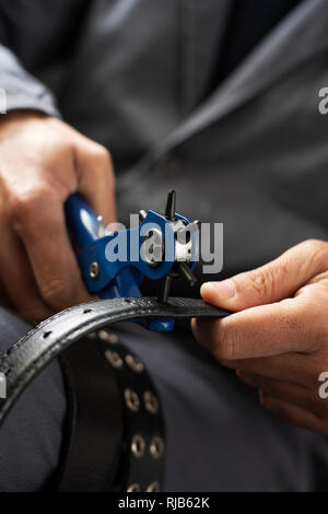 Libre d'un jeune travailleur, homme de race blanche portant un manteau de travail gris, faisant un trou dans une ceinture avec un poinçon en cuir Banque D'Images