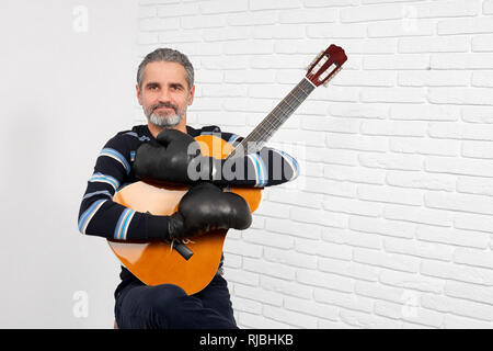 Cheerful man smiling, looking at camera, holding guitare acoustique dans les mains. Homme barbu d'âge mûr vêtu de noir des gants de boxe. Musicien assis en studio et poser. Banque D'Images