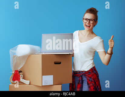 Smiling femme moderne en blanc T-shirt près de boîte en carton montrant ordinateur portable écran blanc et Thumbs up sur fond bleu Banque D'Images