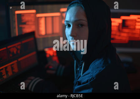 Fille dans le capot à la recherche dans l'appareil photo. Les attaques de pirates et la fraude en ligne sur le fond de l'écran dans l'obscurité. Le concept de sécurité cybernétique Banque D'Images
