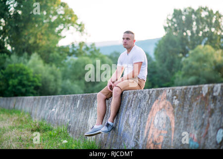 Jeune homme seul assis sur un mur de pierre en été la nature ensoleillée. Banque D'Images