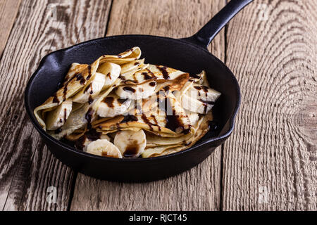 Des crêpes faites maison avec la banane et chocolat topping pour le petit-déjeuner dans la poêle en fonte sur table en bois rustique, Close up, selective focus Banque D'Images