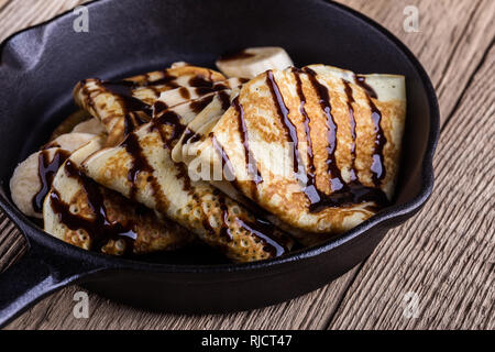 Des crêpes faites maison avec la banane et chocolat topping pour le petit-déjeuner dans la poêle en fonte sur table en bois rustique, Close up, selective focus Banque D'Images