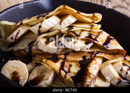 Des crêpes faites maison avec la banane et chocolat topping pour le petit-déjeuner dans la poêle en fonte sur table en bois rustique, Close up, selective focus Banque D'Images