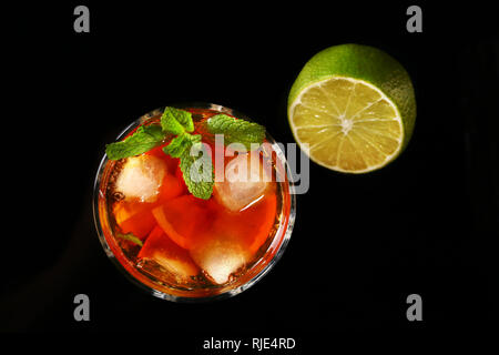 Verre de cocktail rhum foncé avec de la chaux, l'orange, des cubes de glace et de feuilles de menthe sur fond miroir noir. Vue d'en haut. Banque D'Images