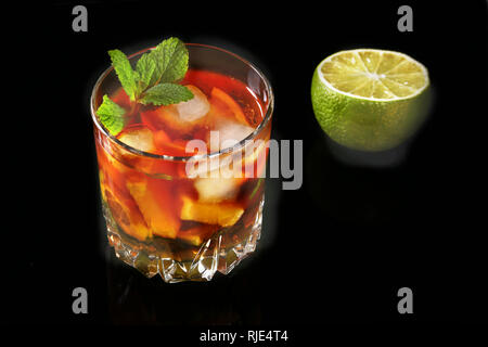 Verre de cocktail rhum foncé avec de la chaux, l'orange, des cubes de glace et de feuilles de menthe sur fond miroir noir. Banque D'Images