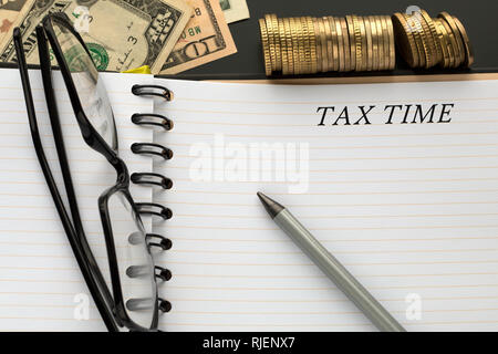 Bloc-notes avec temps d'impôts mots, crayon, verres et billets en dollars, des piles de pièces de monnaie sur l'arrière-plan. Banque D'Images