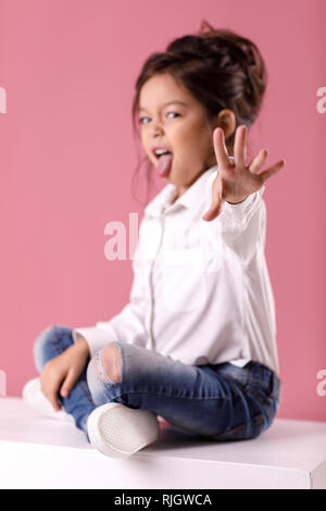 Adorable petit enfant fille en chemise blanche avec hairstyle montrant langue sur fond rose. Les émotions humaines et l'expression faciale Banque D'Images